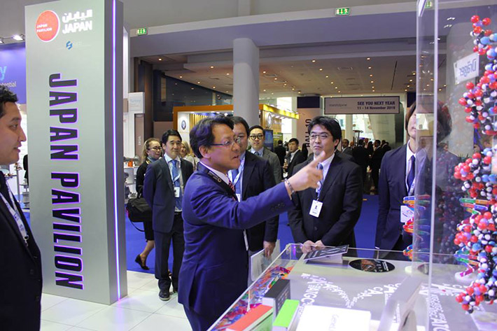 日本パビリオンに展示されたDNA二重らせん模型の前で経産省の関副大臣（左）に最先端ゲノムテクノロジーについて説明する相澤准教授（中央）