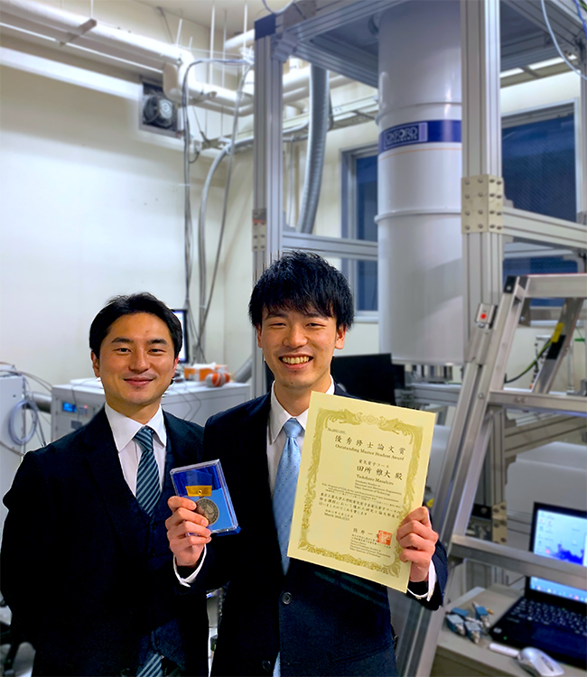 Masahiro Tadokoro (right) and Associate Professor Tetsuo Kodera (left). The equipment at the back right is the cryogenic refrigerator used in the research.
