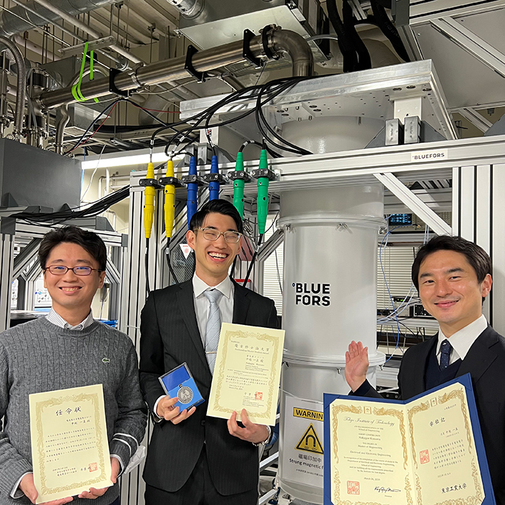小寺哲夫准教授（右）、中越一真さん（真ん中）、米田淳特任准教授（左）。奥は実験で用いた希釈冷凍機。